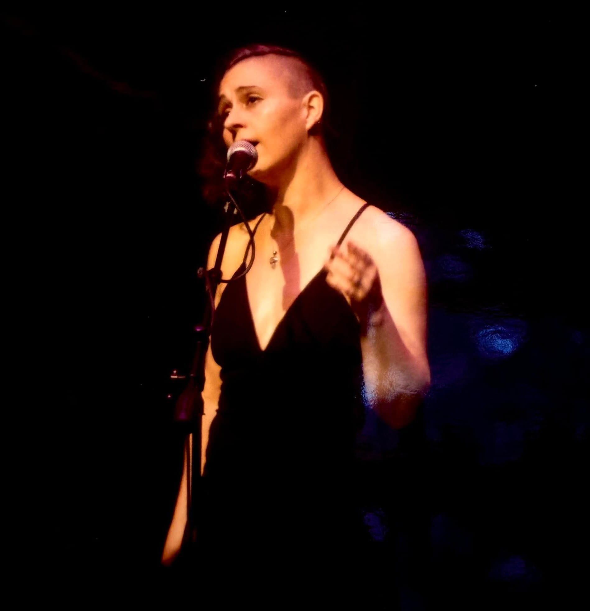Singer in a black dress performs at a microphone under warm stage lighting.