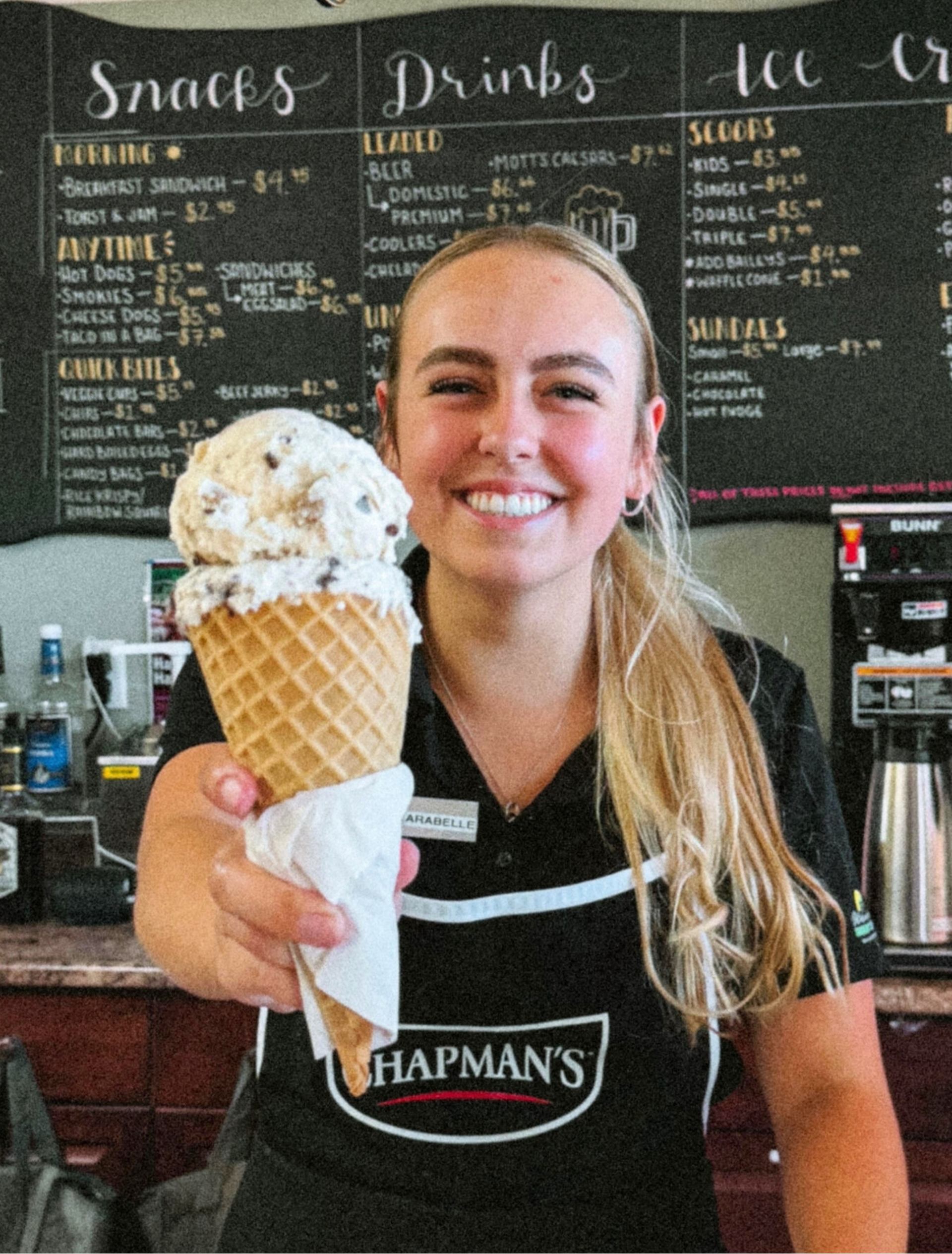 Ice cream shop worker holding waffle cone in front of chalkboard menu.