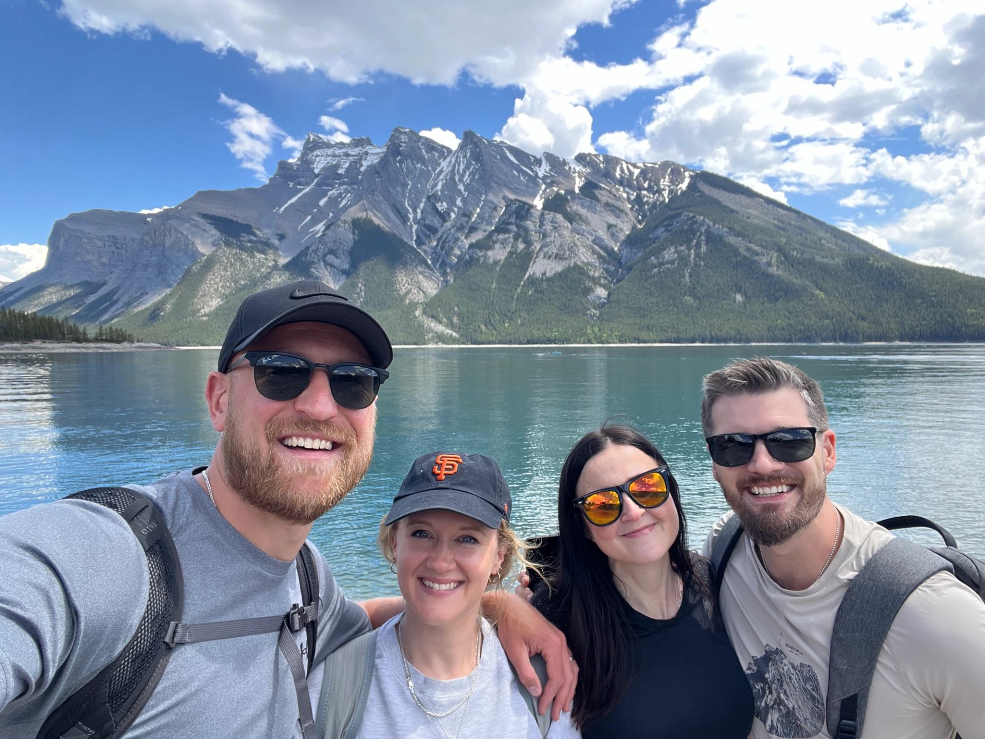 Four friends in sunglasses pose by a calm mountain lake with rocky peaks.