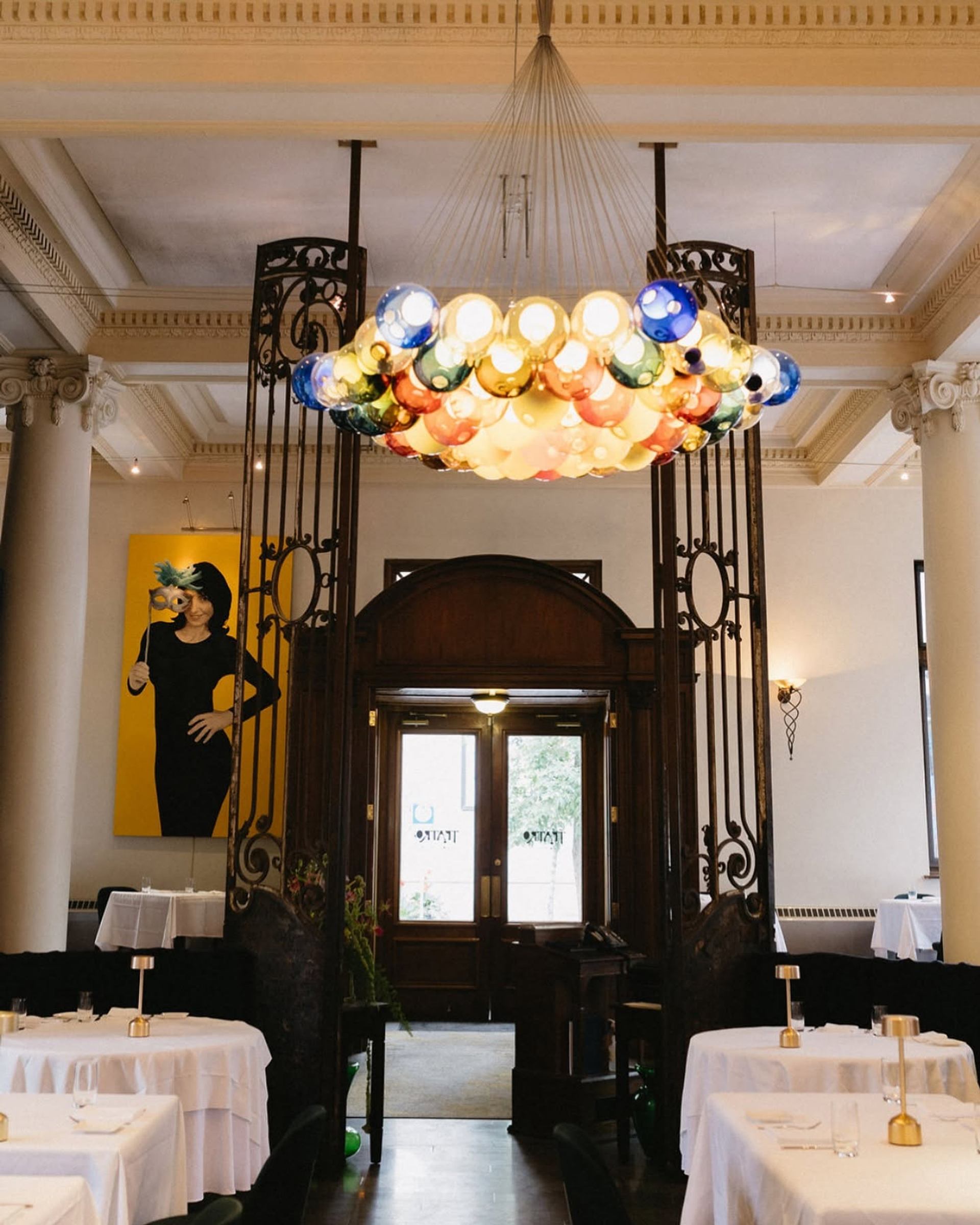 Dining room with colourful glass chandelier, white tablecloths and large portrait on yellow wall.