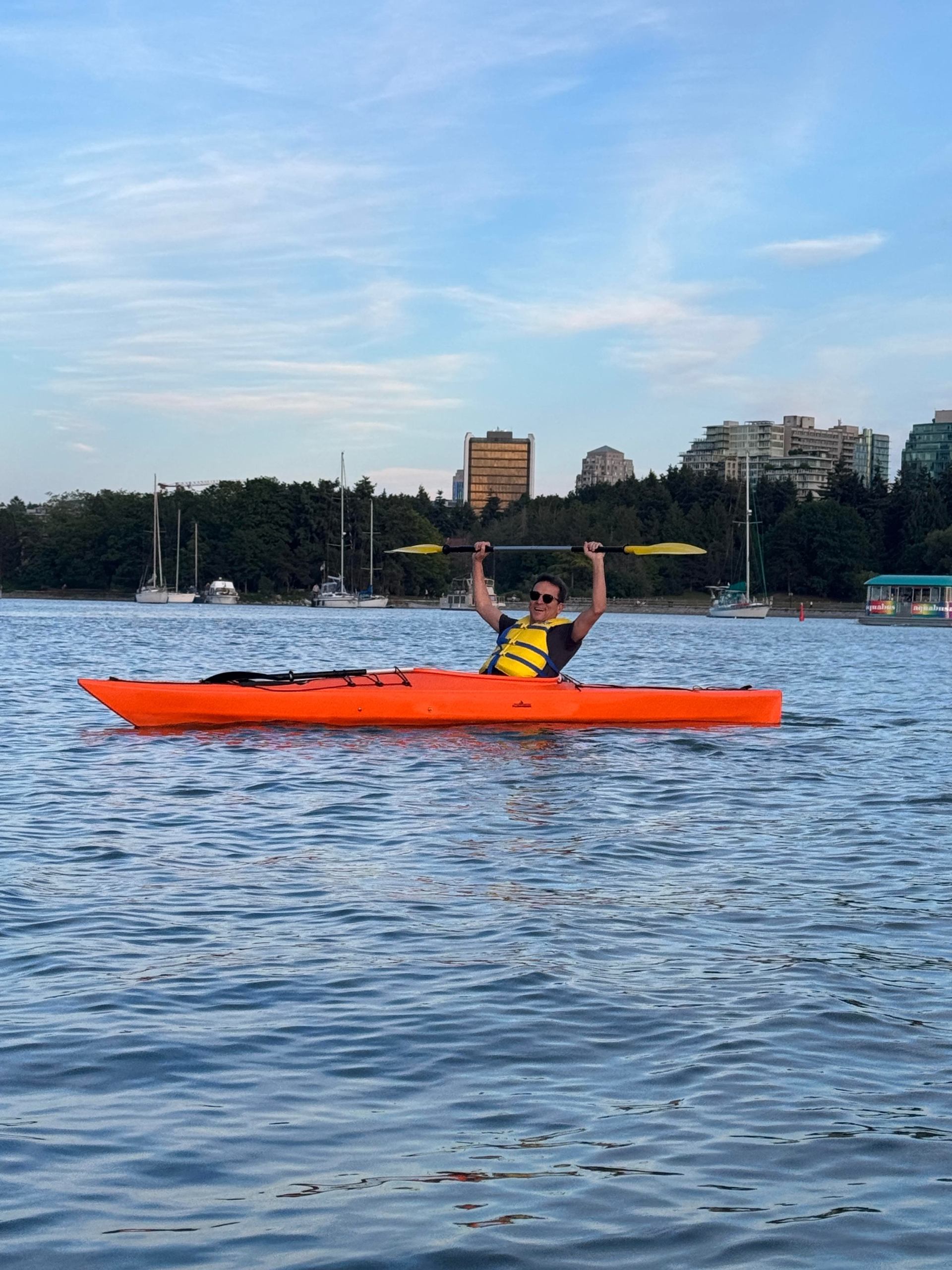 Person in yellow life jacket raises paddle while sitting in orange kayak on harbour.