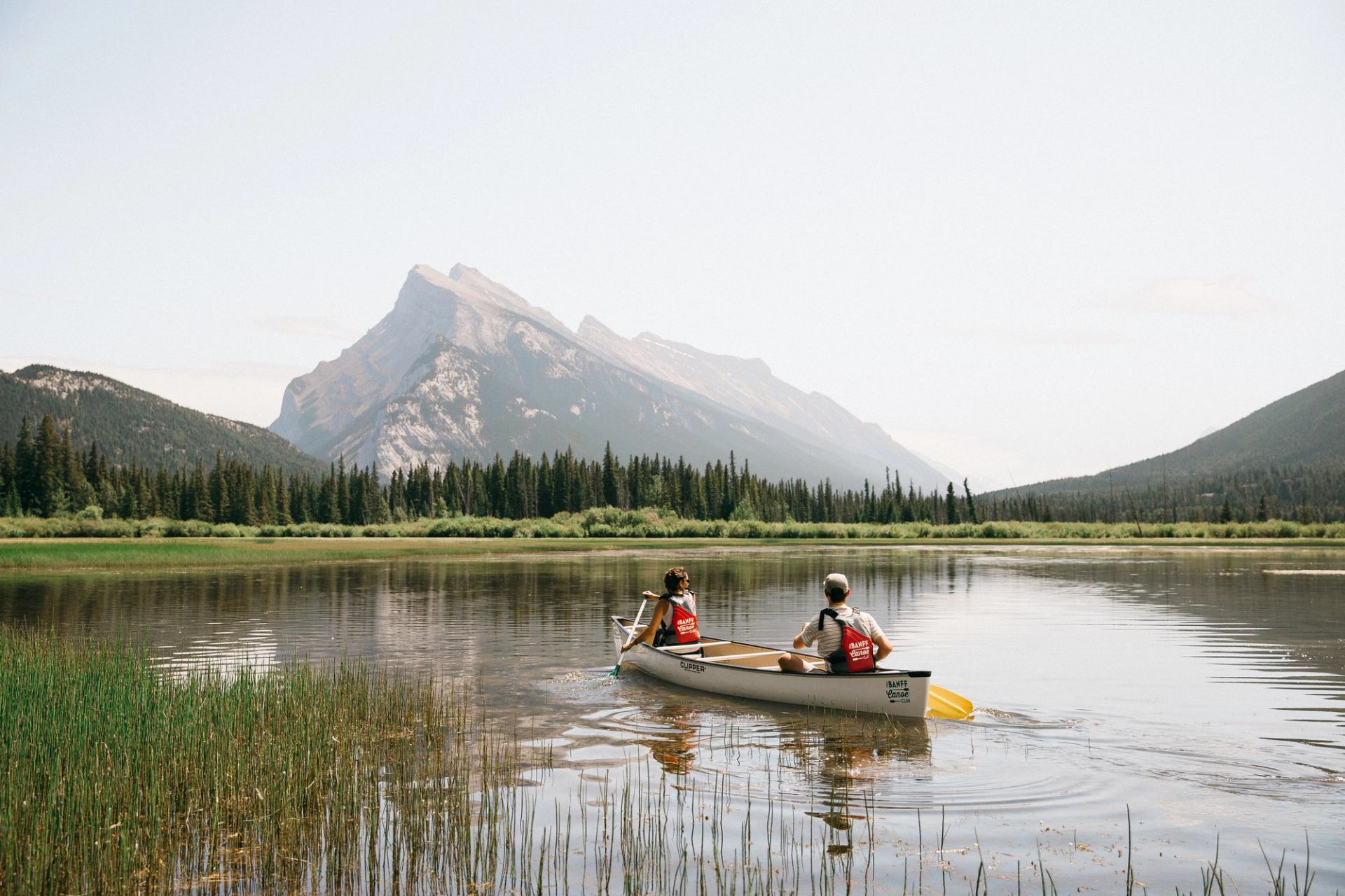 Two people canoeing on a calm lake with mountains and forest in the background.