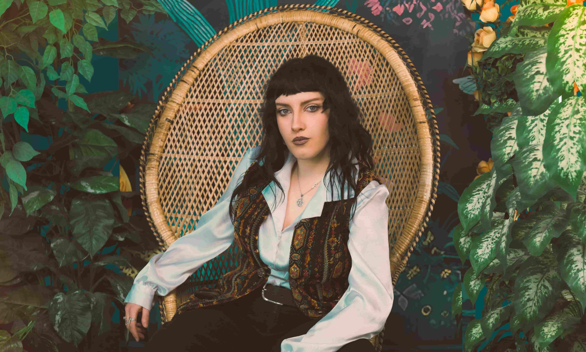 Person with dark hair sits in wicker chair surrounded by lush indoor plants.