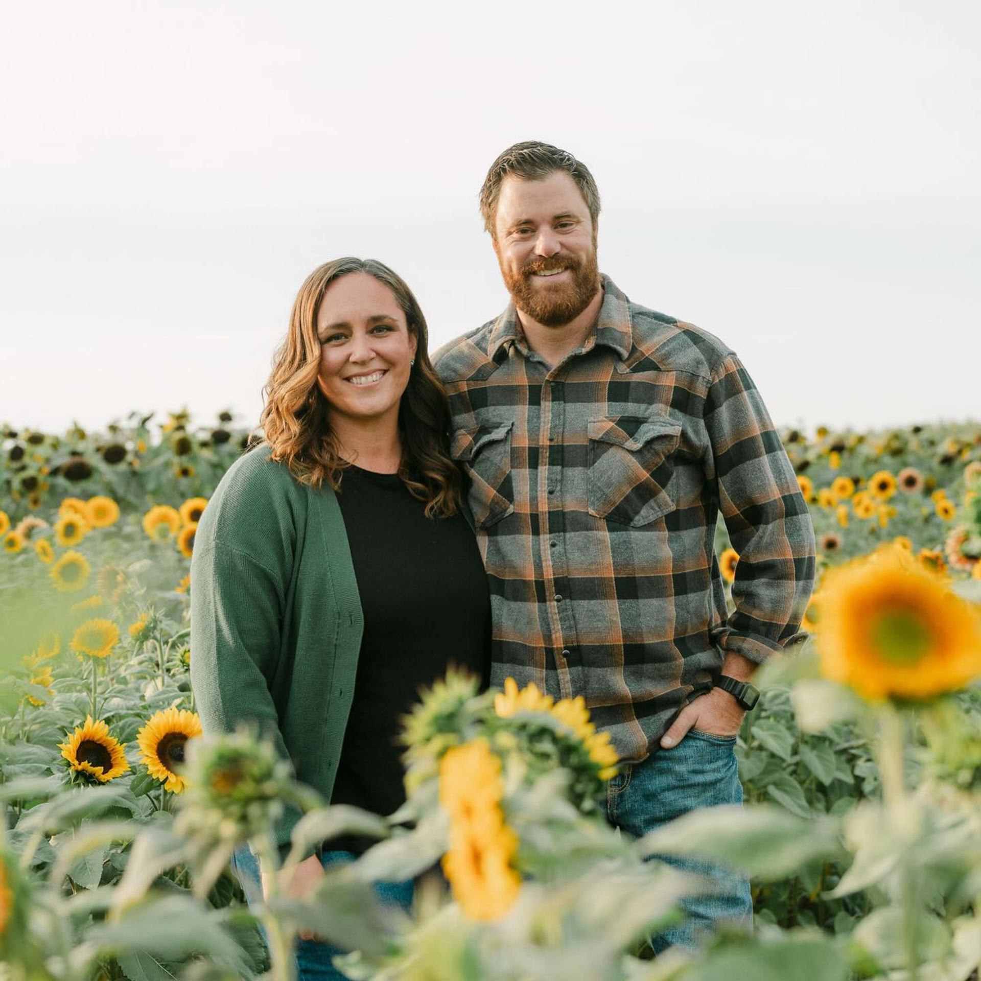 Two adults stand close together in a sunflower field, facing the camera.
