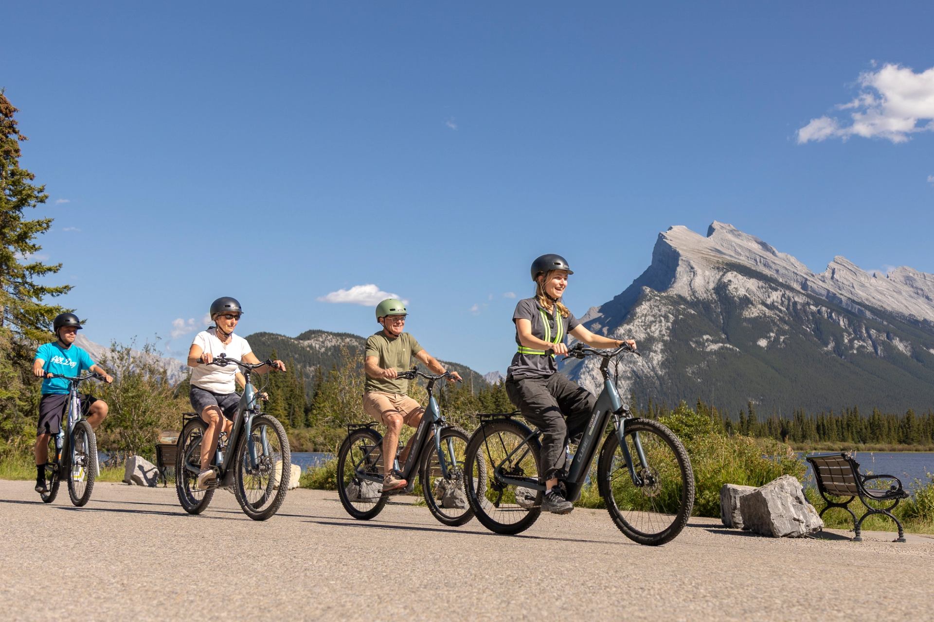 Cyclists ride e-bikes on paved path beside lake, with forested mountains behind.
