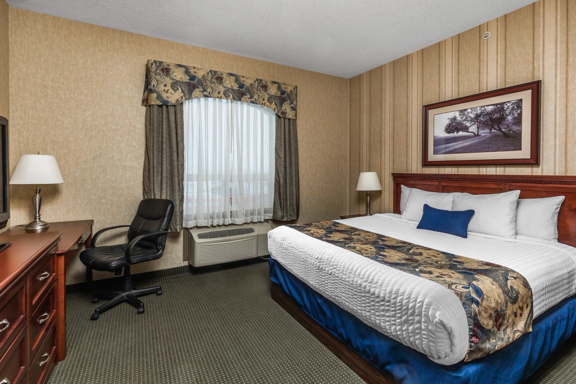 Hotel bedroom with double bed, desk chair, lamps, and framed landscape wall art.