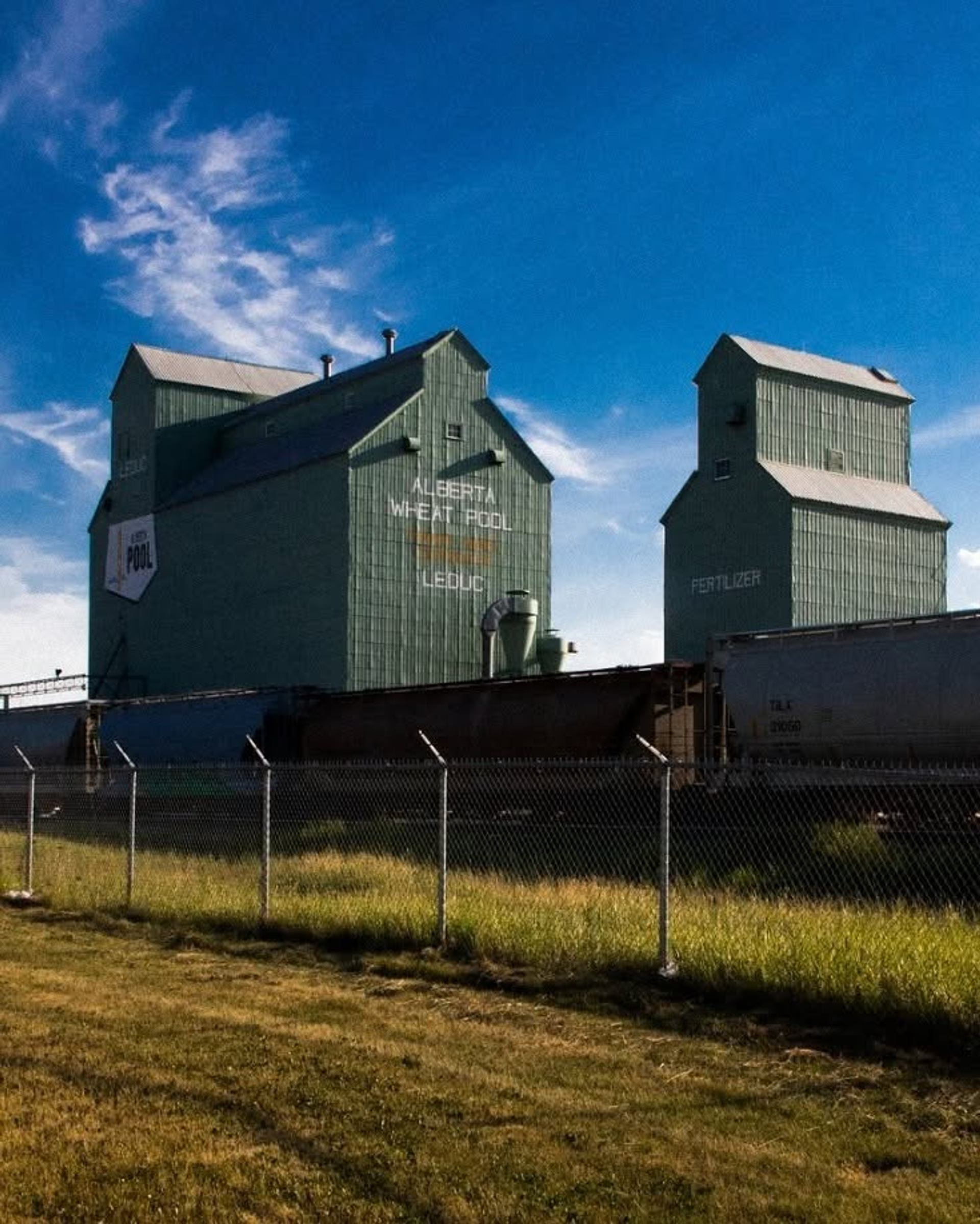 Two historic Alberta Wheat Pool grain elevators stand beside rail cars under a bright blue prairie sky.