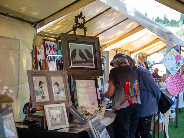 Art vendor at outdoor market booth under white tent, framed bird and animal pictures on display.