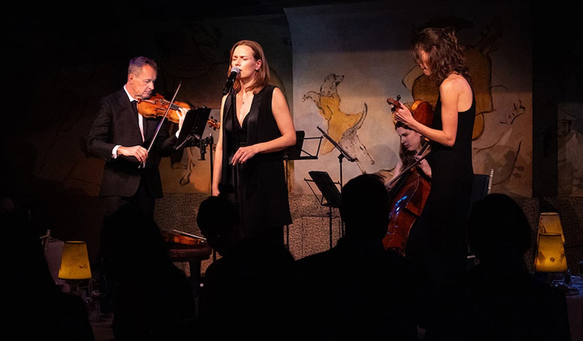 Vocalist and chamber ensemble performing live on an intimate stage with violin, cello, and warm theatrical lighting.