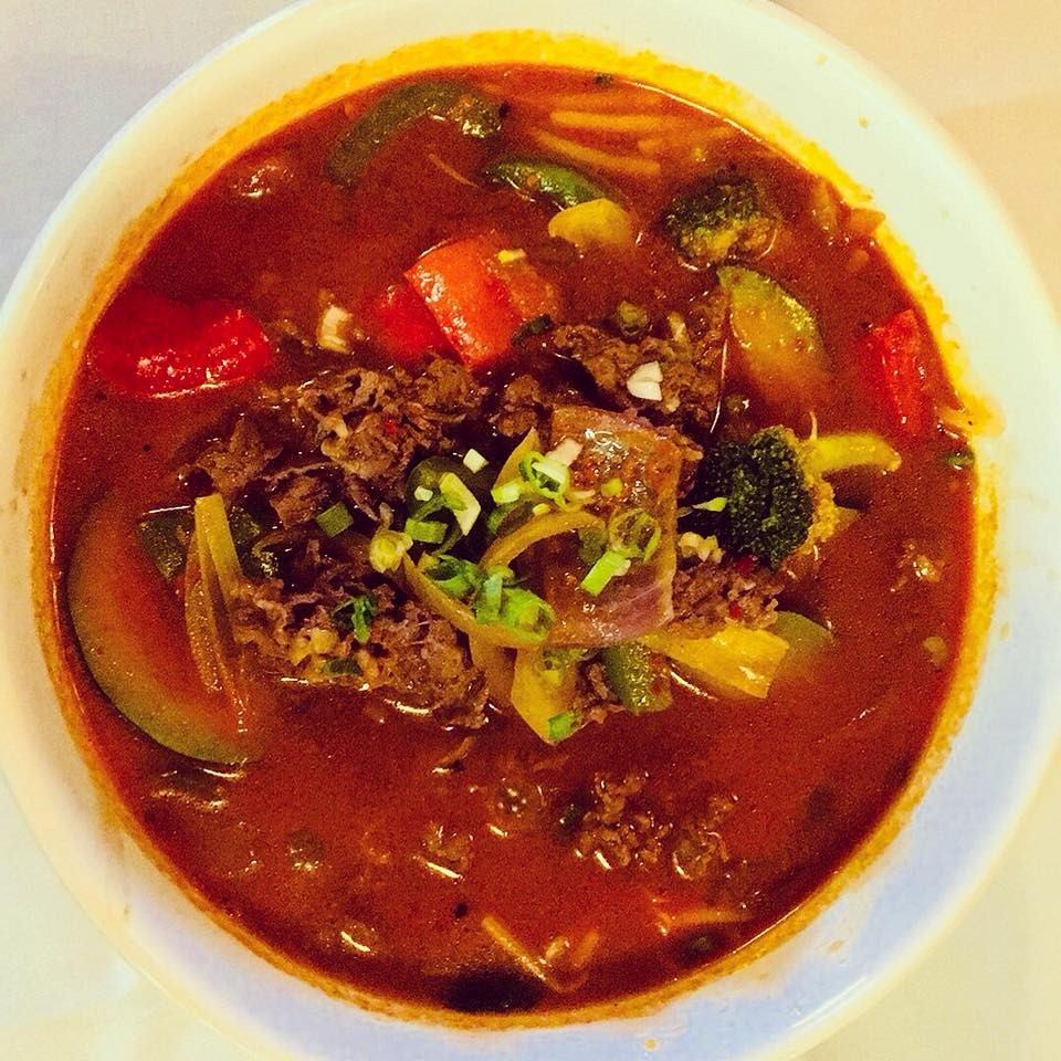 Bowl of red broth soup with meat, veggies, and fresh herb garnish.