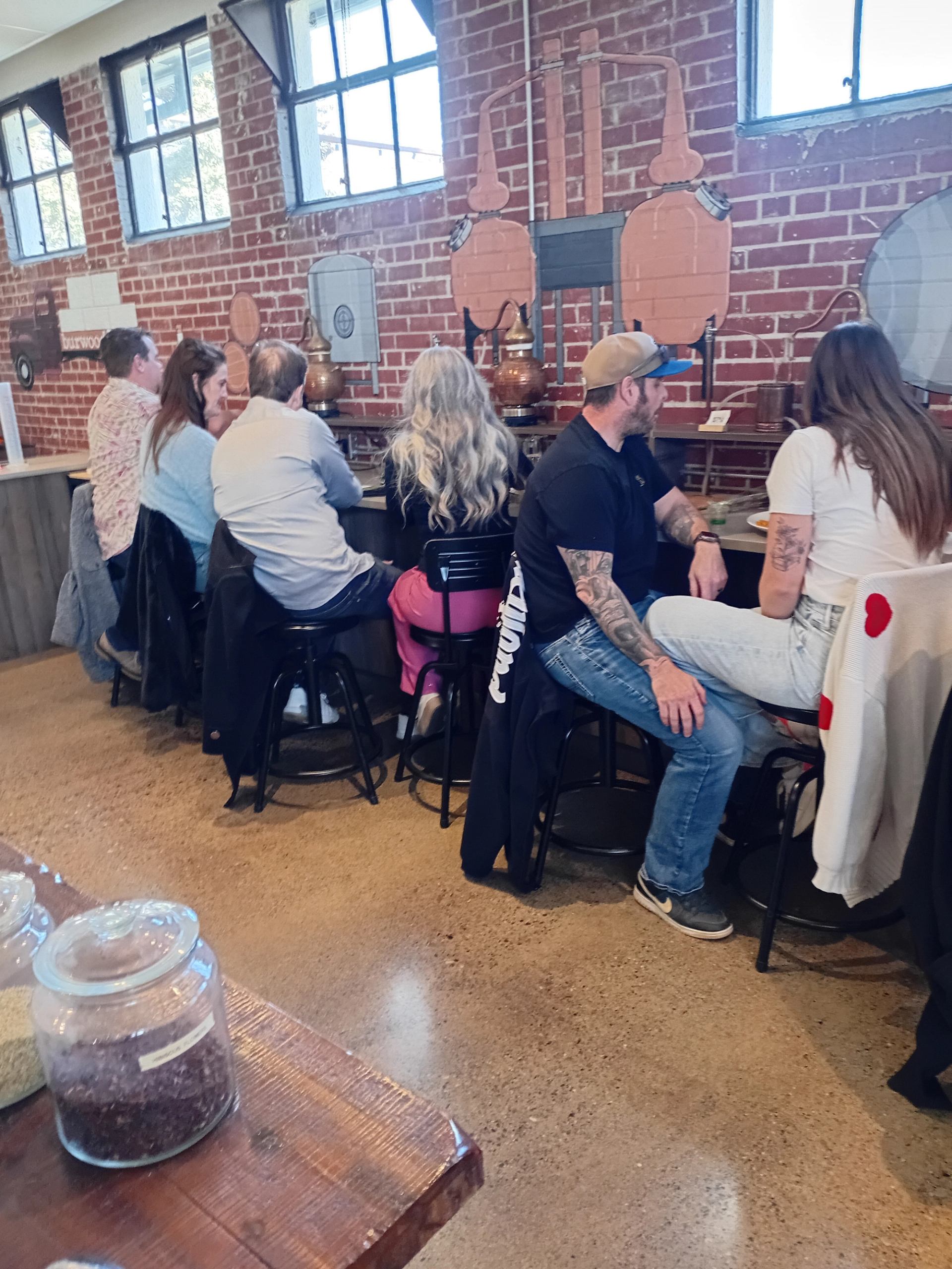 Visitors sit at a counter beneath copper distilling equipment and brick walls.