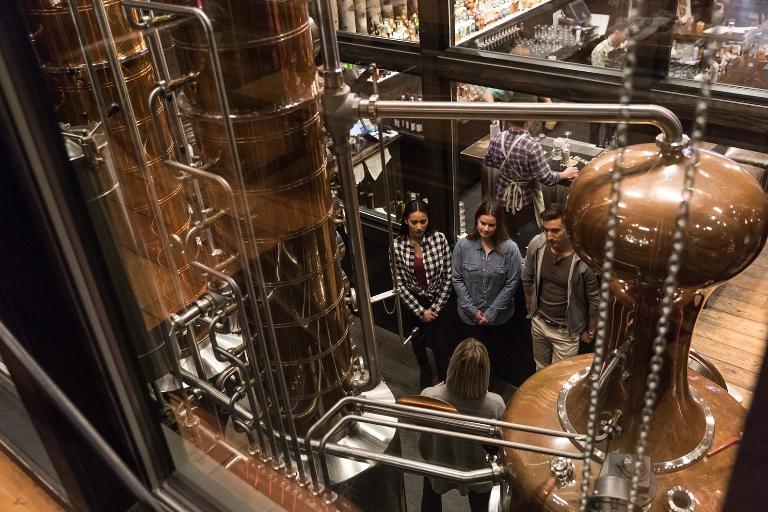 A group of people observing large copper distilling equipment in a distillery, with a bar in the background.