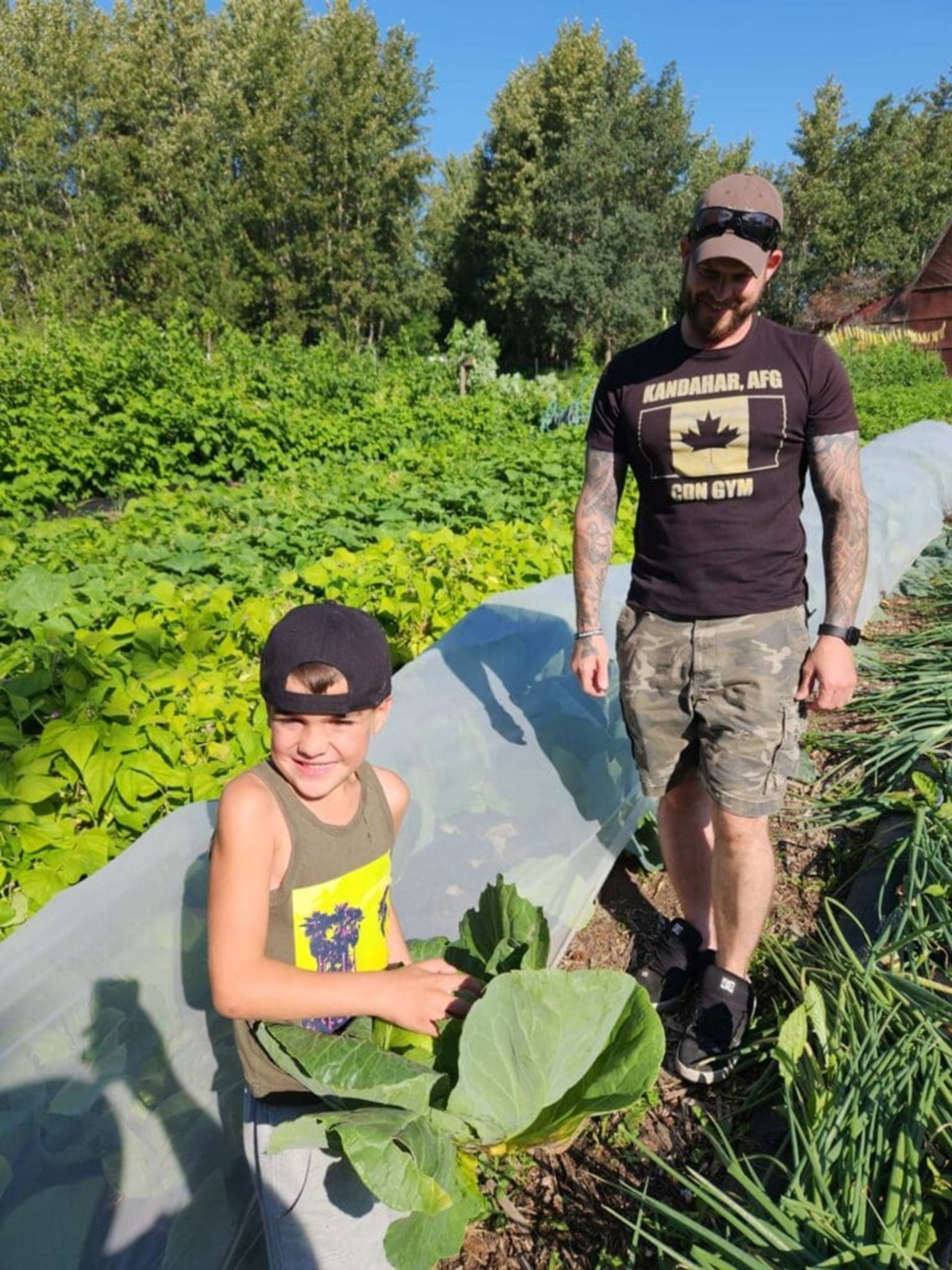 Child and adult stand in vegetable garden, holding large leafy cabbage near plants.
