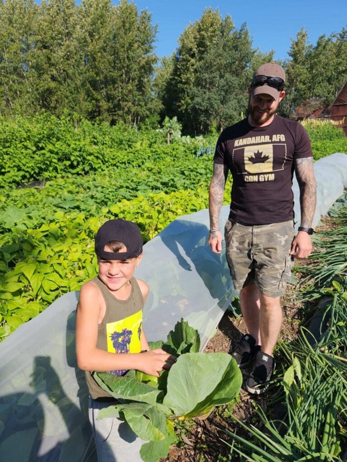 Child and adult stand in vegetable garden, holding large leafy cabbage near plants.