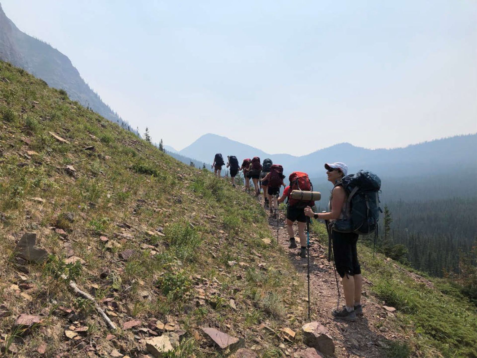 Backpackers hiking uphill on a narrow trail along a mountainside with distant ridgelines.