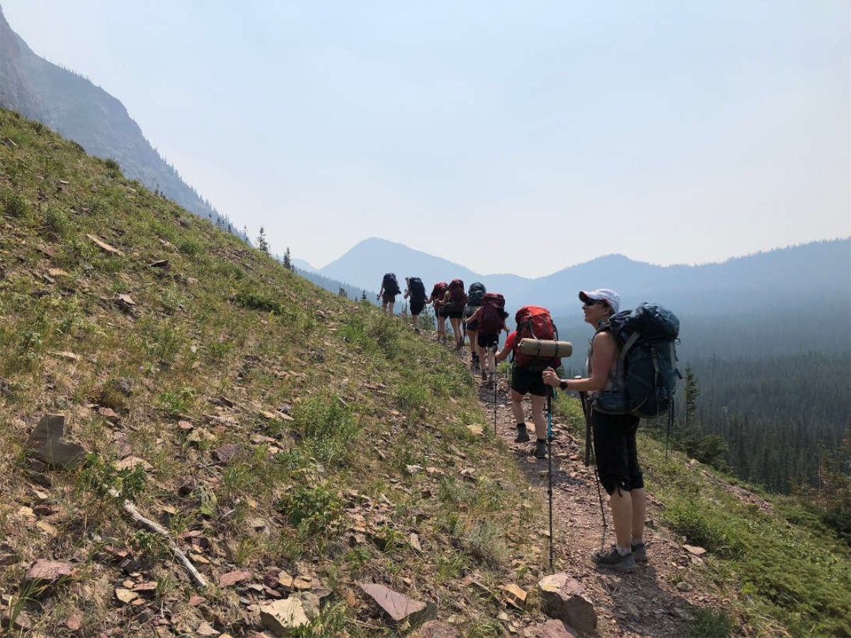 Backpackers hiking uphill on a narrow trail along a mountainside with distant ridgelines.