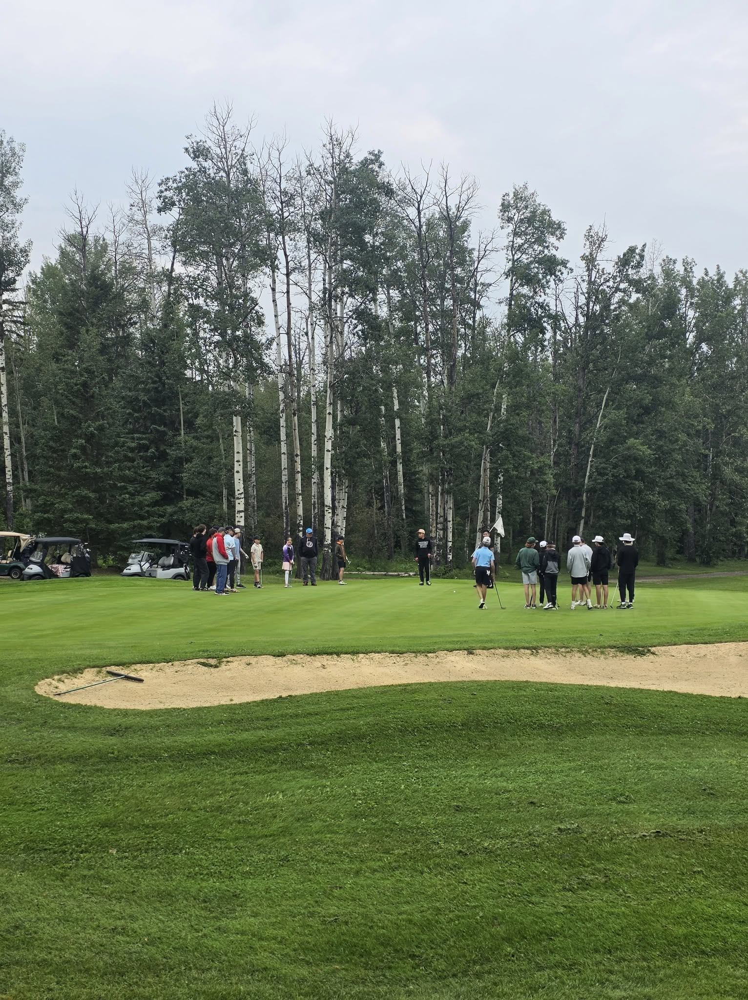 Golfers gather on a putting green beside a sand bunker and wooded area.