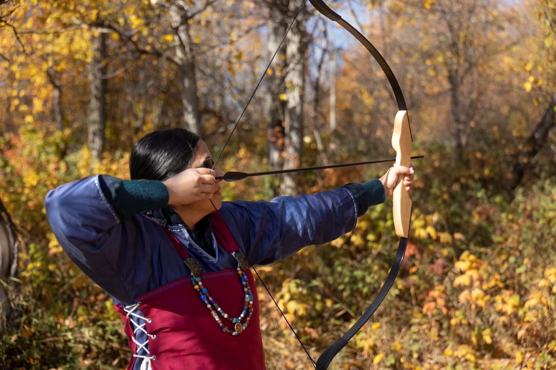 A person in traditional clothing aims a bow and arrow in an autumn forest.