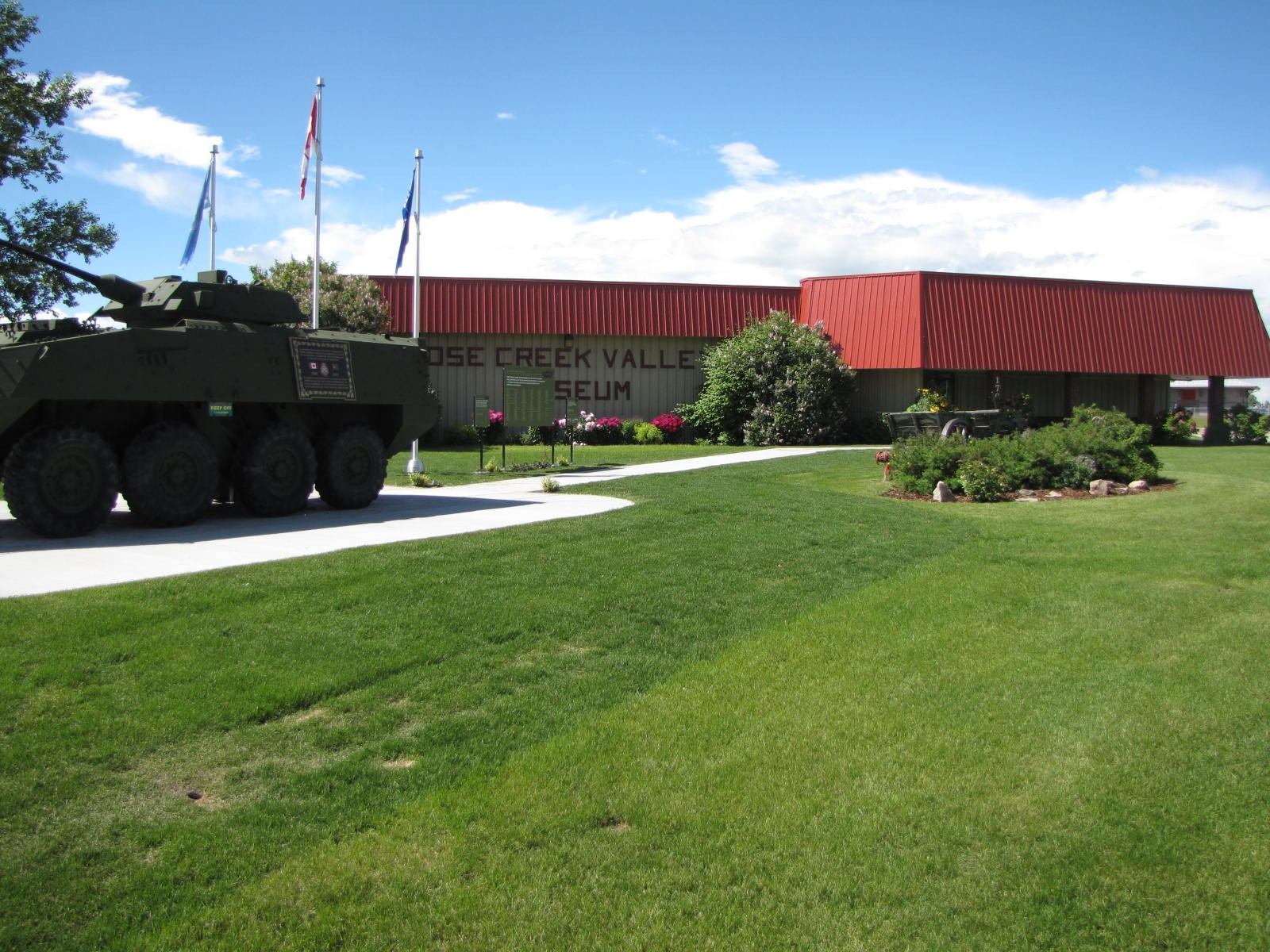 Exterior of the Nose Creek Valley Museum with a military vehicle, flags, and landscaped lawn in front.