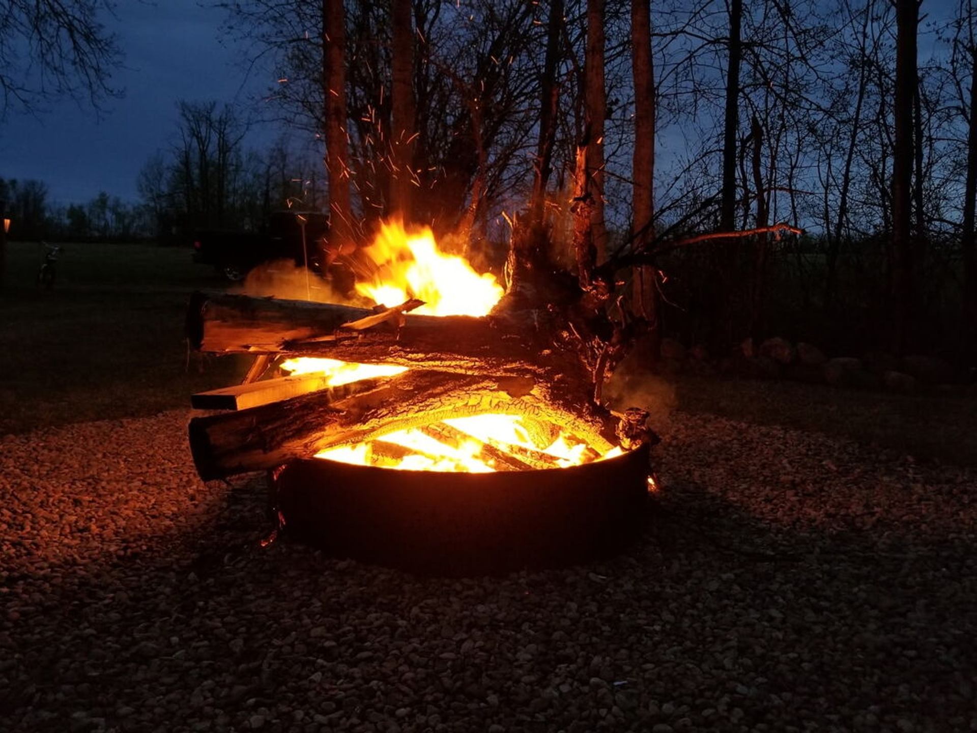 Large campfire in metal ring with tall flames and sparks among trees at dusk.