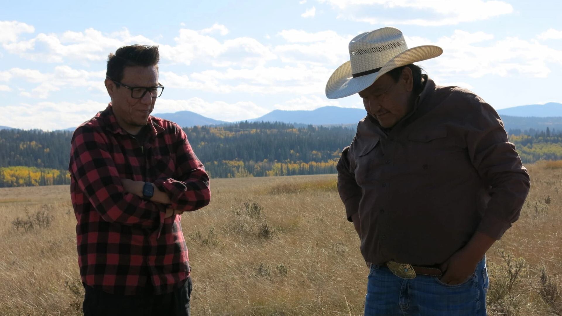 Two men stand in a grassy field with mountains and fall trees in the background.