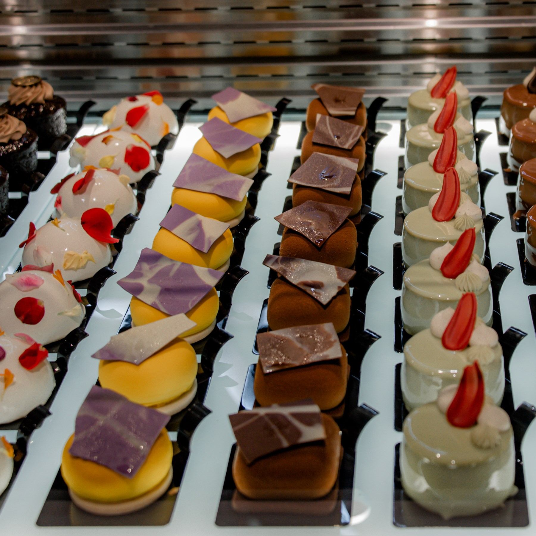 Assorted pastries in bakery display trays, including glazed cakes, chocolate squares, and cream dess.