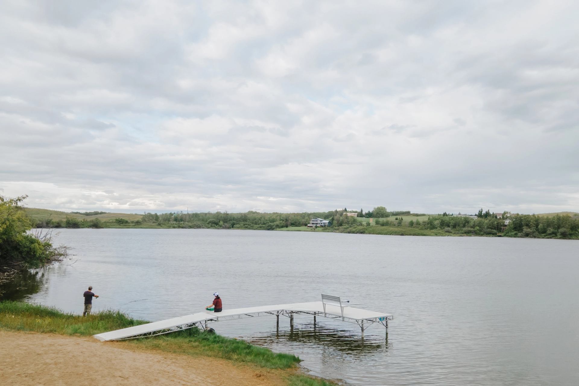 Lakeside dock with seated person, another standing on shore, distant houses and trees.