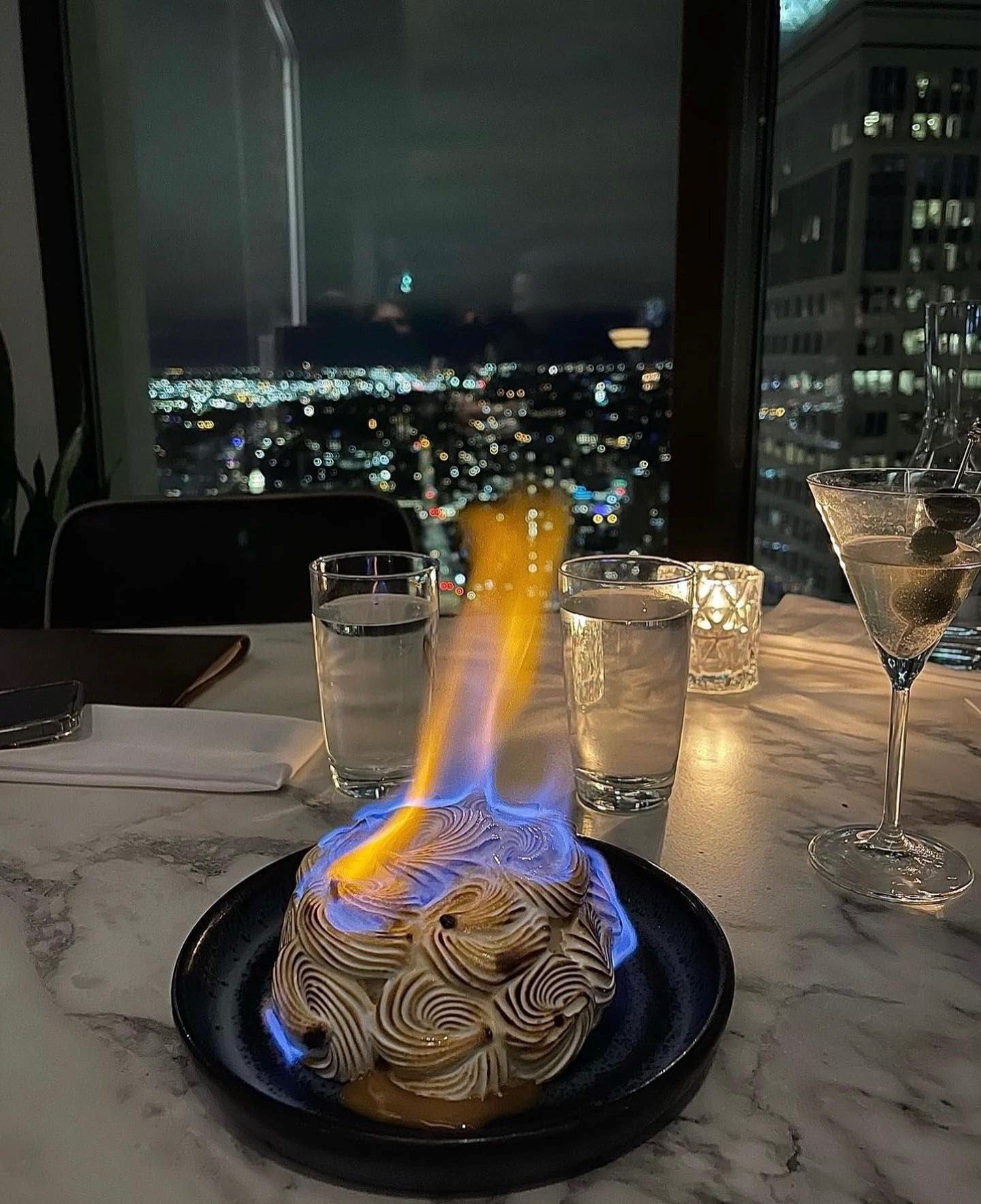 Flaming Baked Alaska dessert on a marble table with city night view through a window in background.