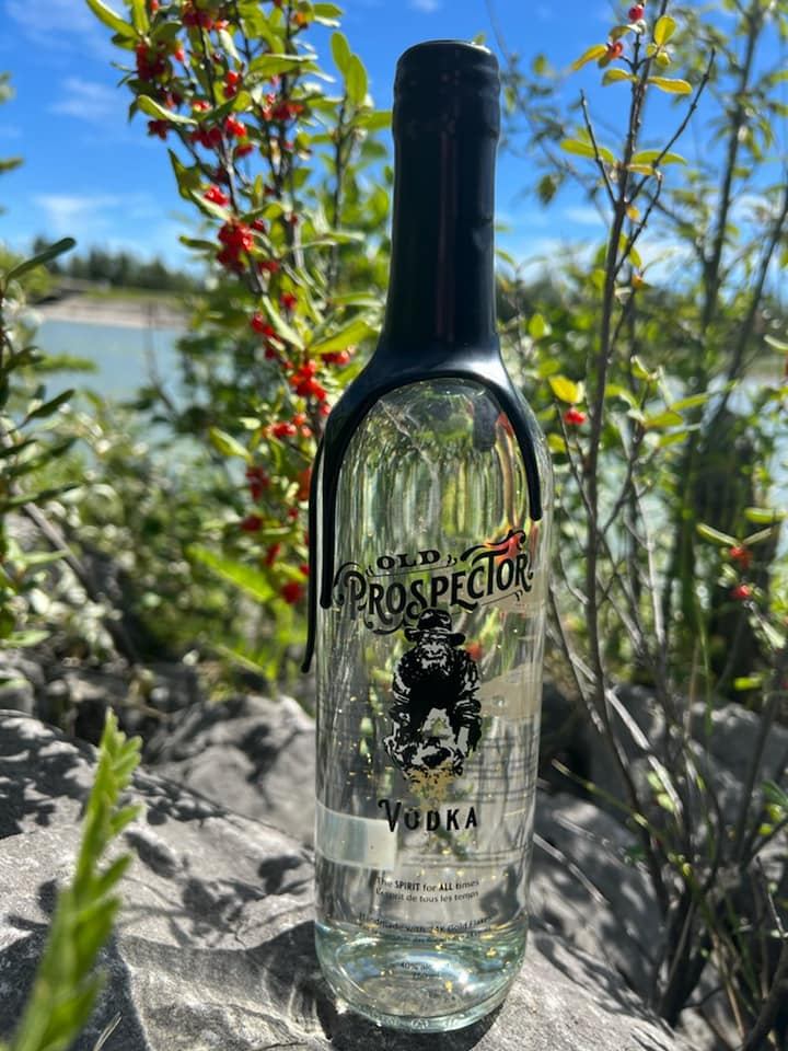 Old Prospector Vodka bottle with gold flakes on a rock, set against a backdrop of red-berried bushes, water, and blue sky.
