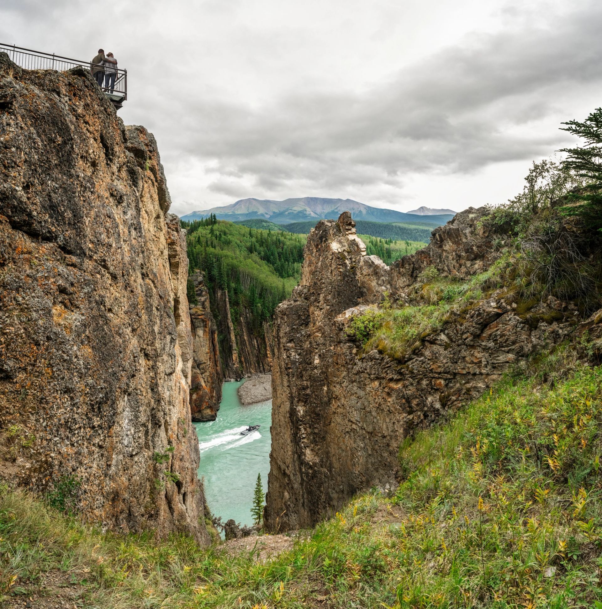 Visitors overlook a turquoise river winding through a rocky canyon beneath cloudy mountains.