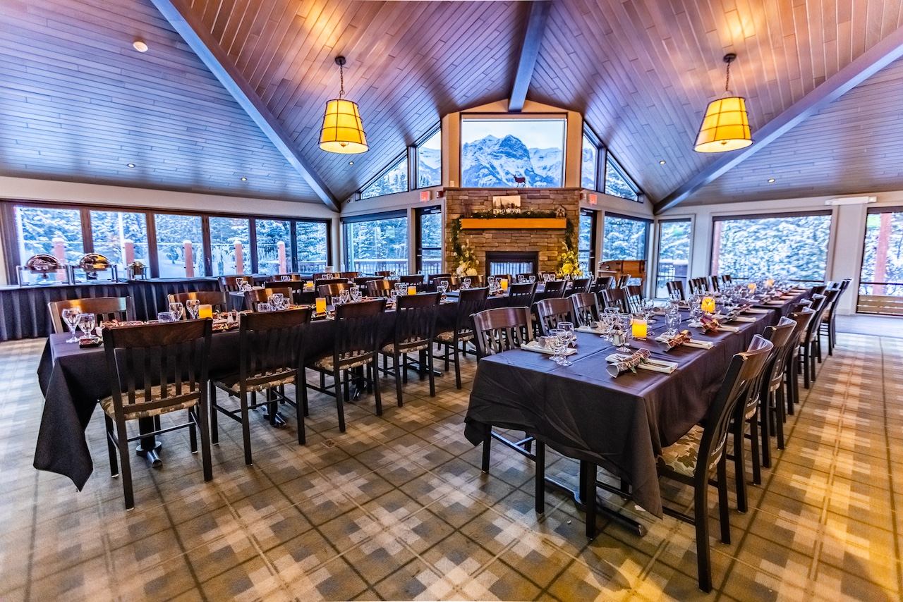 A spacious event room with vaulted wood ceilings, long tables set for dining, and large windows overlooking a snowy landscape. A fireplace features a mountain image projected above it.