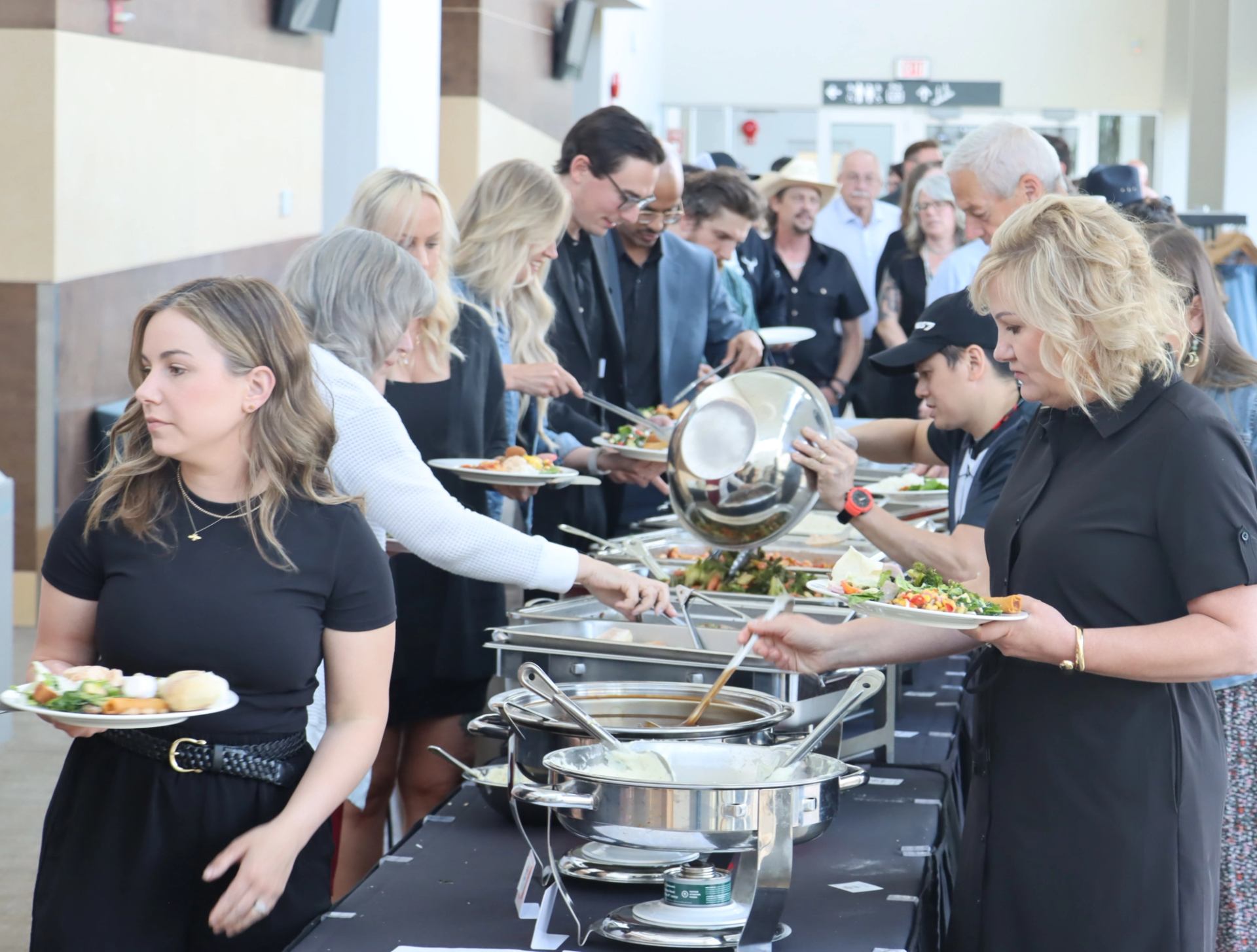 People serve themselves food from buffet chafing dishes at a crowded indoor event.
