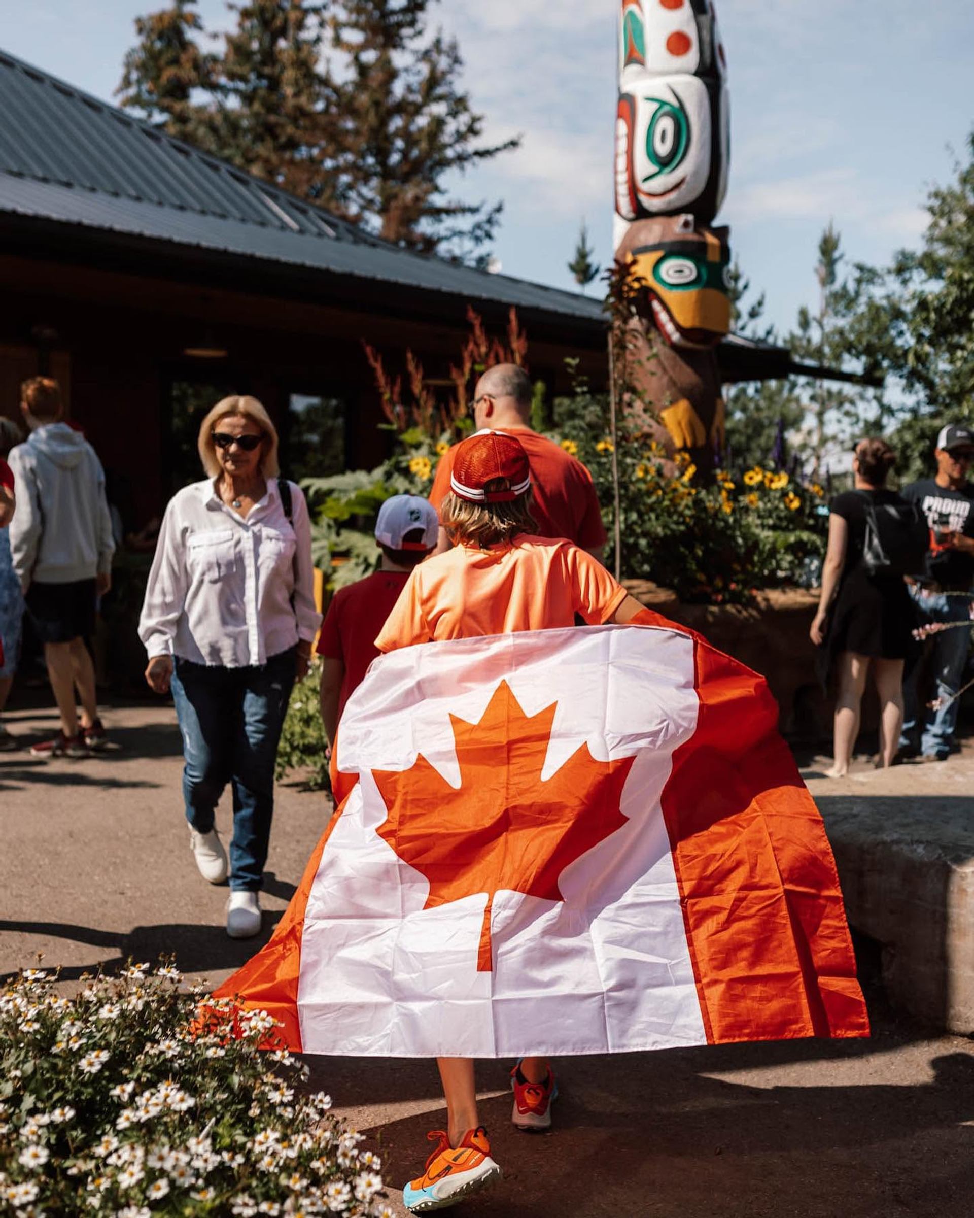 Person wearing a Canadian flag walks near totem pole and garden outside building.