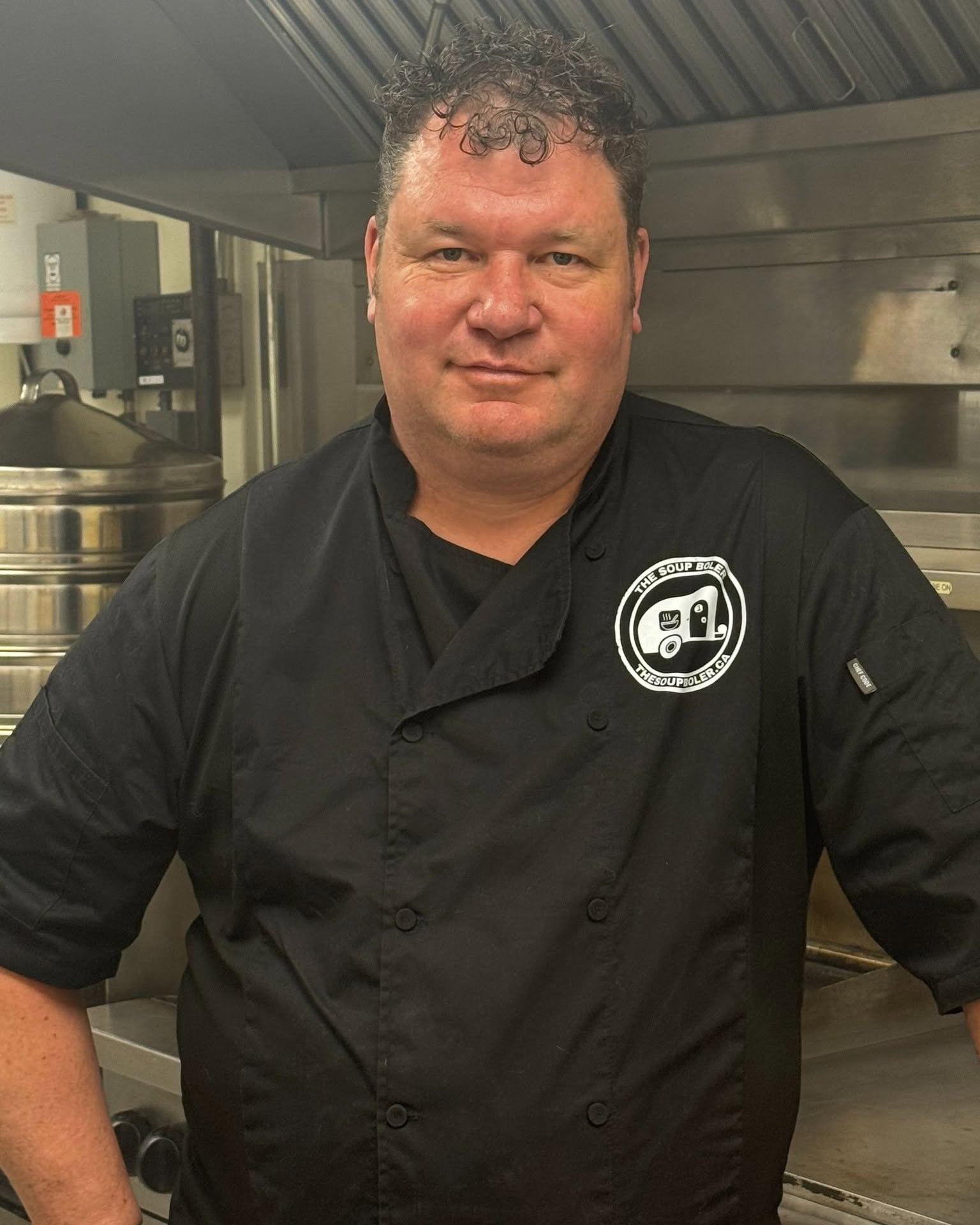 Chef in black uniform with The Soup Boiler logo stands in commercial kitchen.