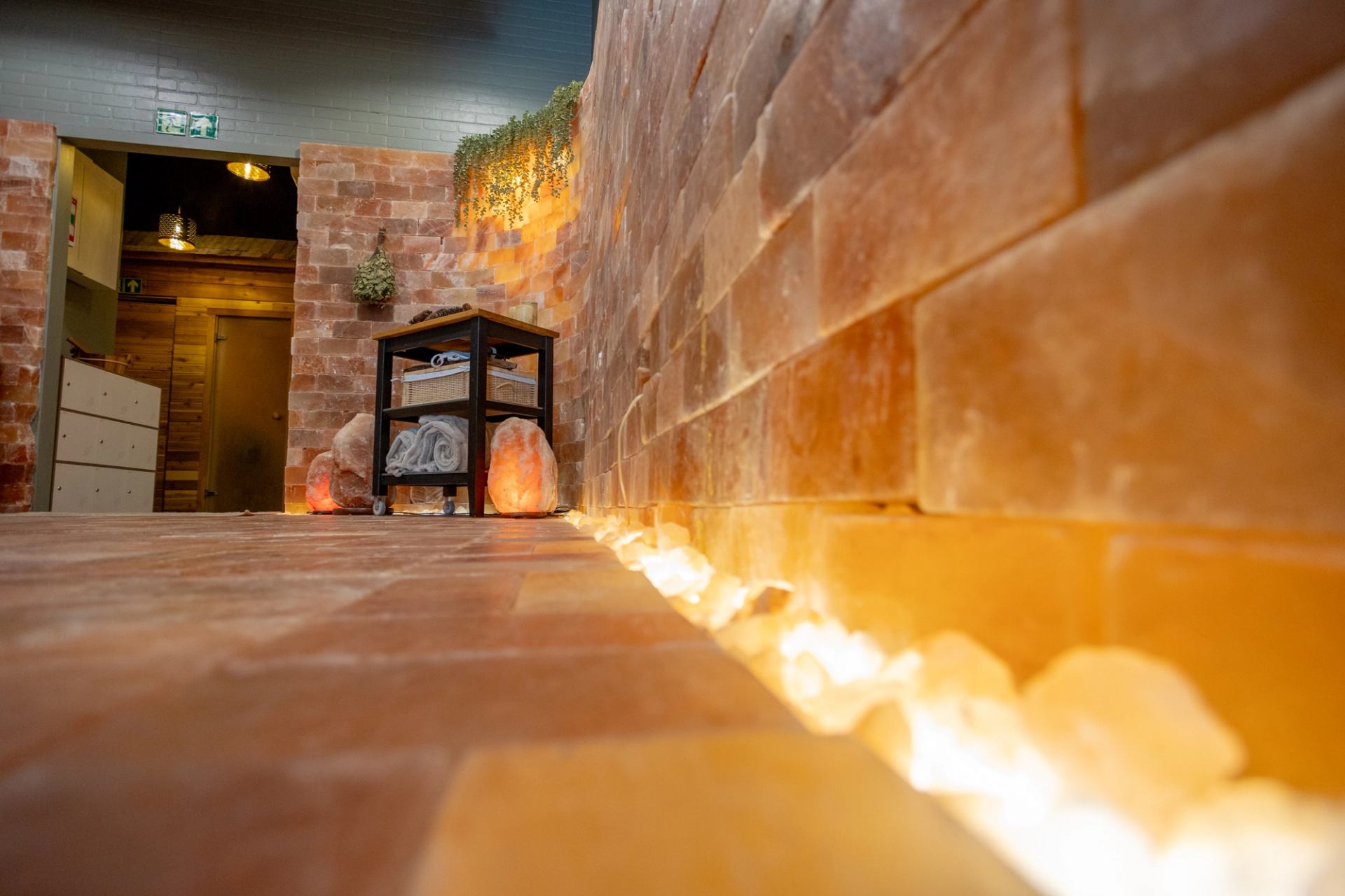 A low-angle shot showcases a glowing, illuminated pathway along a curved wall made entirely of pink Himalayan salt blocks. A small utility cart with towels and decorative salt lamps sits in the background.