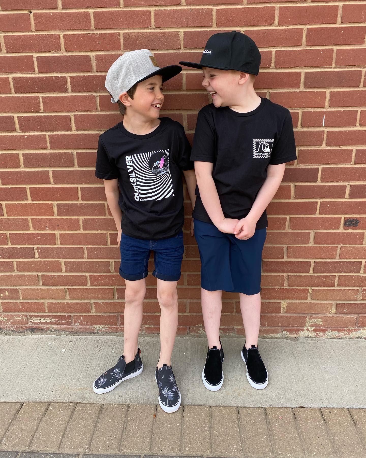 Two children stand outside a brick wall, wearing black T-shirts and sneakers.