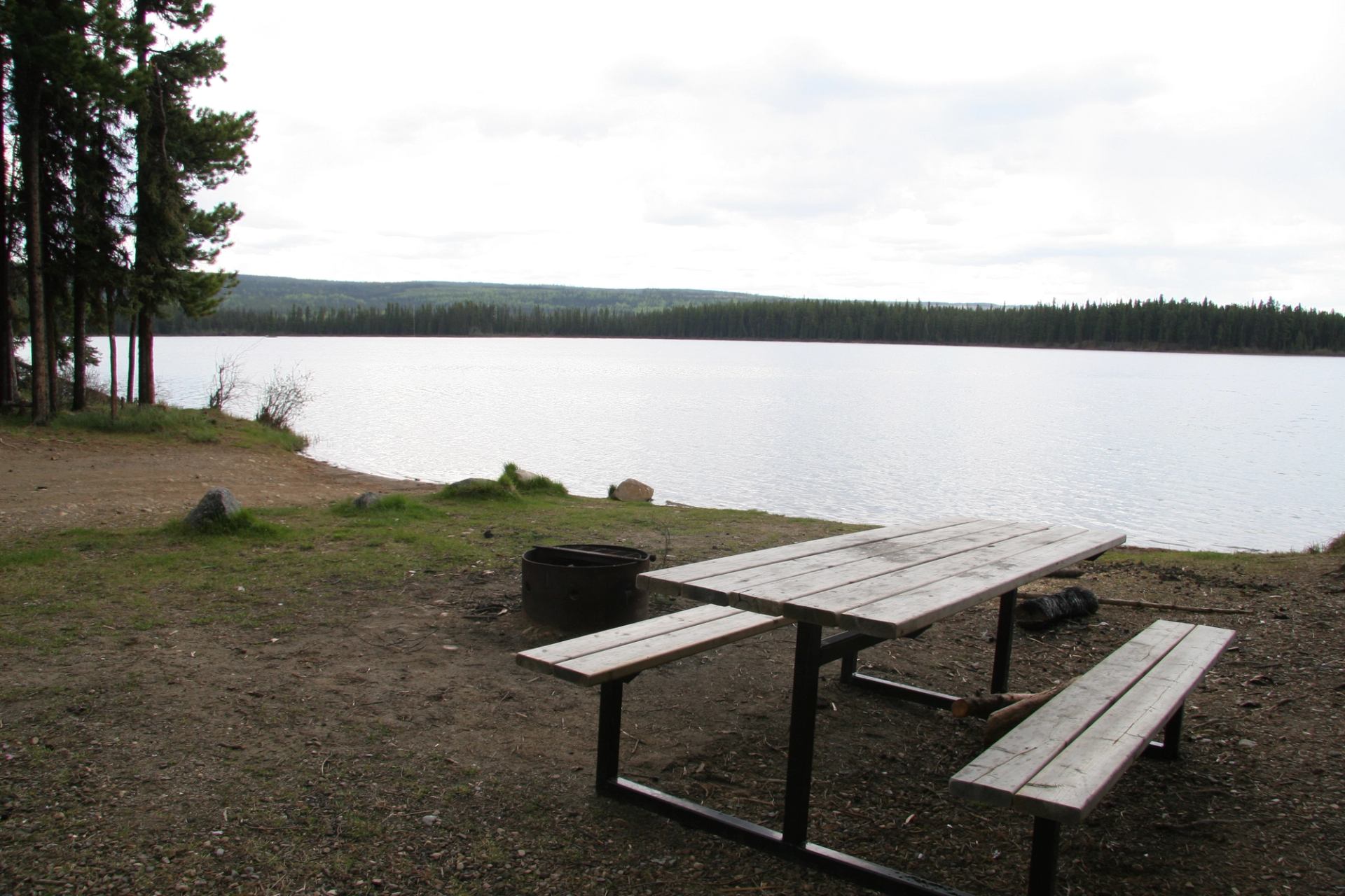 Picnic table and fire pit by calm lake under cloudy sky.