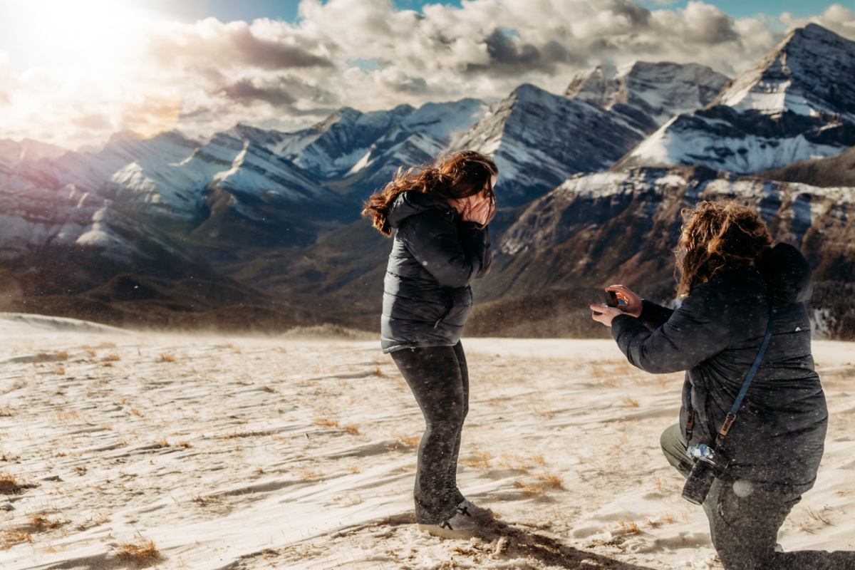 A photographer captures a proposal moment on a windy snowy mountain slope.