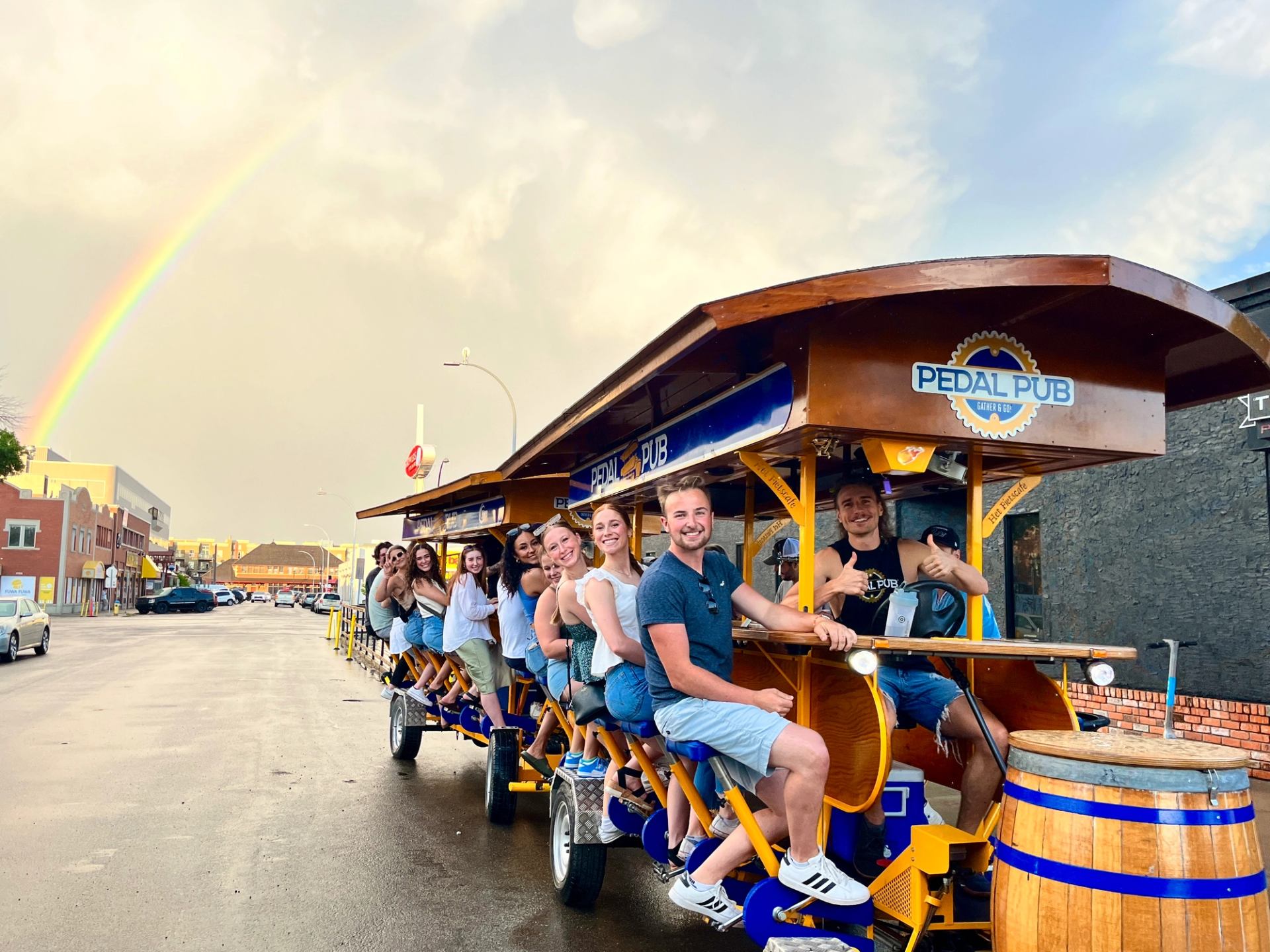 Group riding a Pedal Pub bike along an Edmonton street under a bright rainbow.