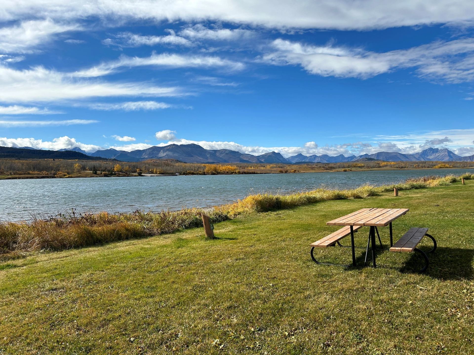 Wooden picnic table on grassy shore beside lake with distant mountain range.