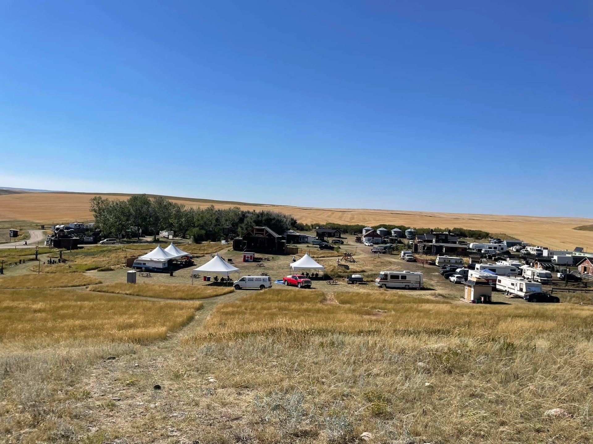 An outdoor event with multiple RVs, white tents, and trees in a golden field under a clear blue sky.