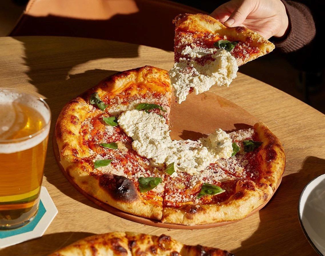 Close-up of a pizza topped with cheese and herbs next to a glass of beer on a wooden table.