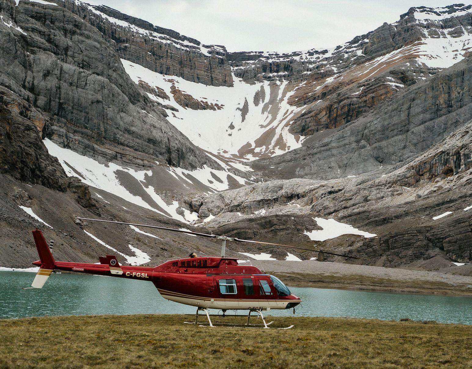 A red helicopter parked beside a turquoise alpine lake surrounded by cliffs.