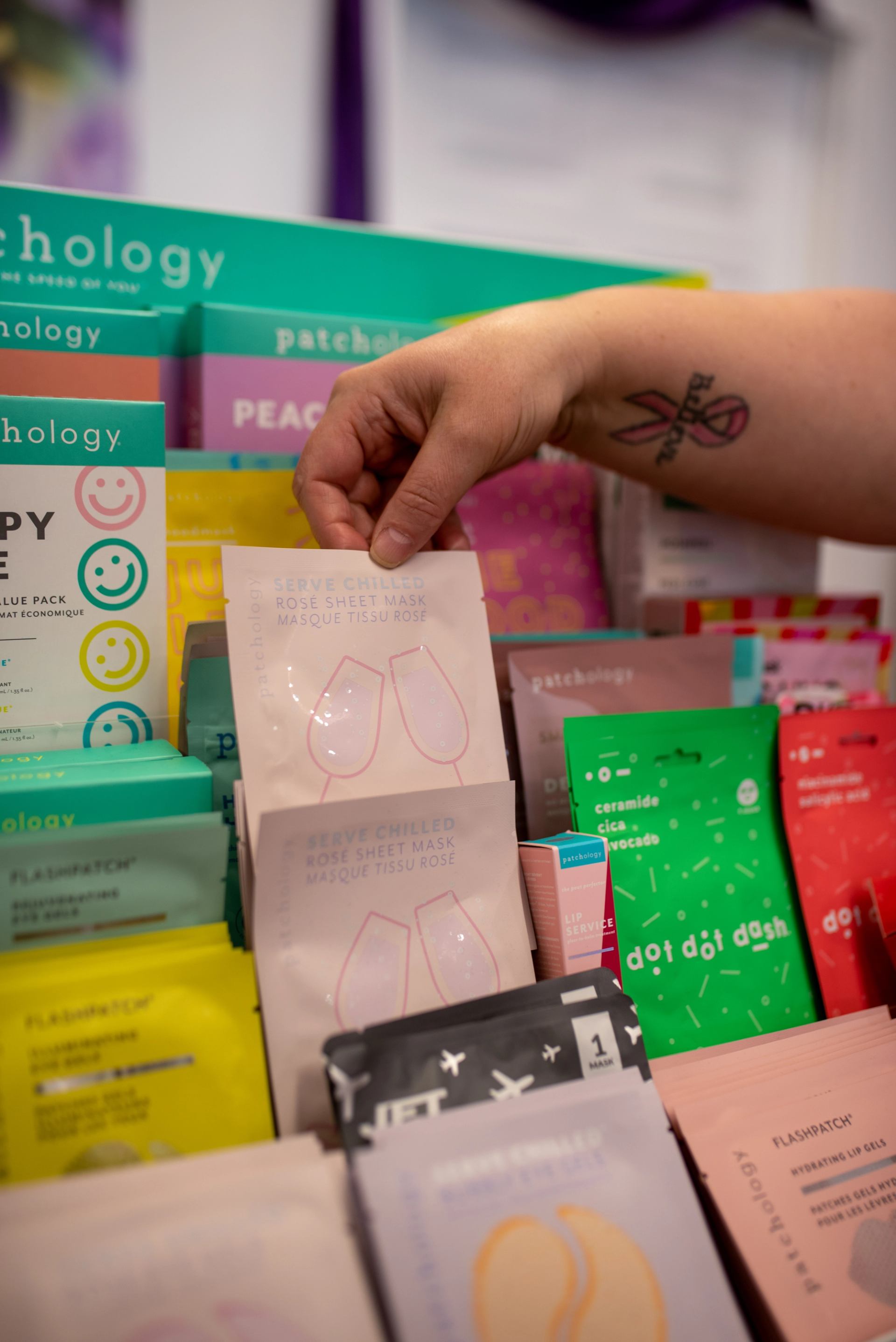 Hand with ribbon tattoo selecting Patchology face mask from colourful display of skincare packets.