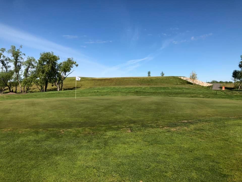 Golf course fairway with flag, green grass, and rolling hills under a blue sky.