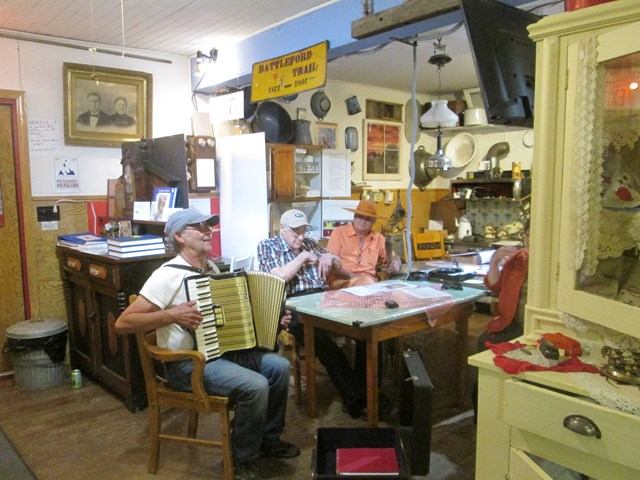 Three people playing music on accordion and fiddle inside a room filled with vintage artifacts.