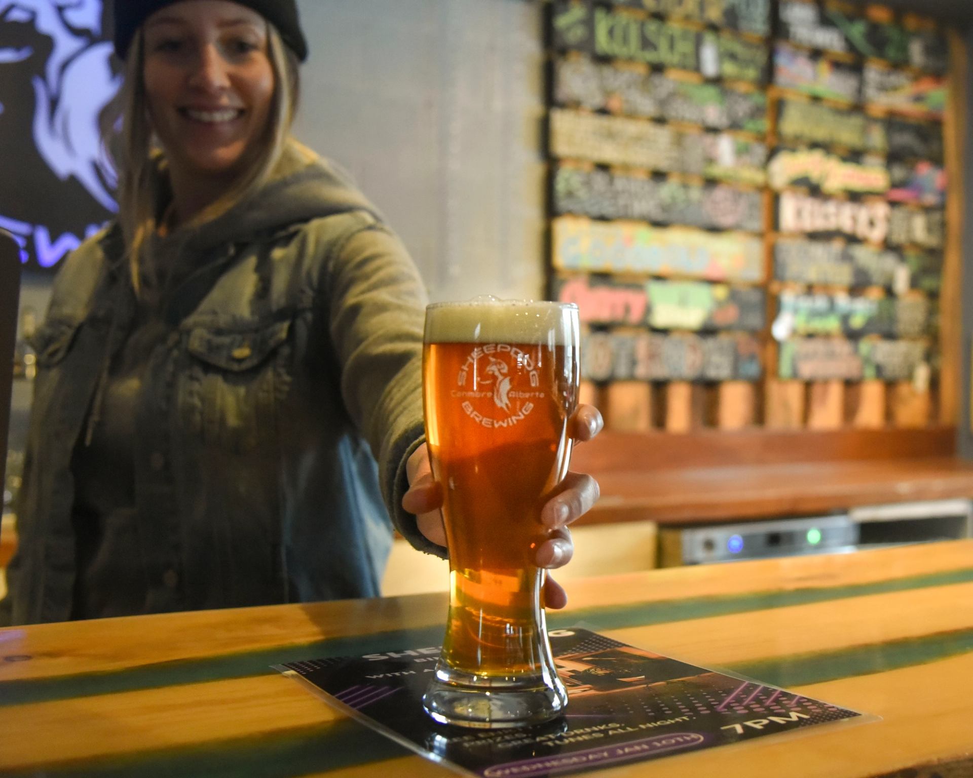 Bartender holds a foamy pint of amber beer across a wooden bar.