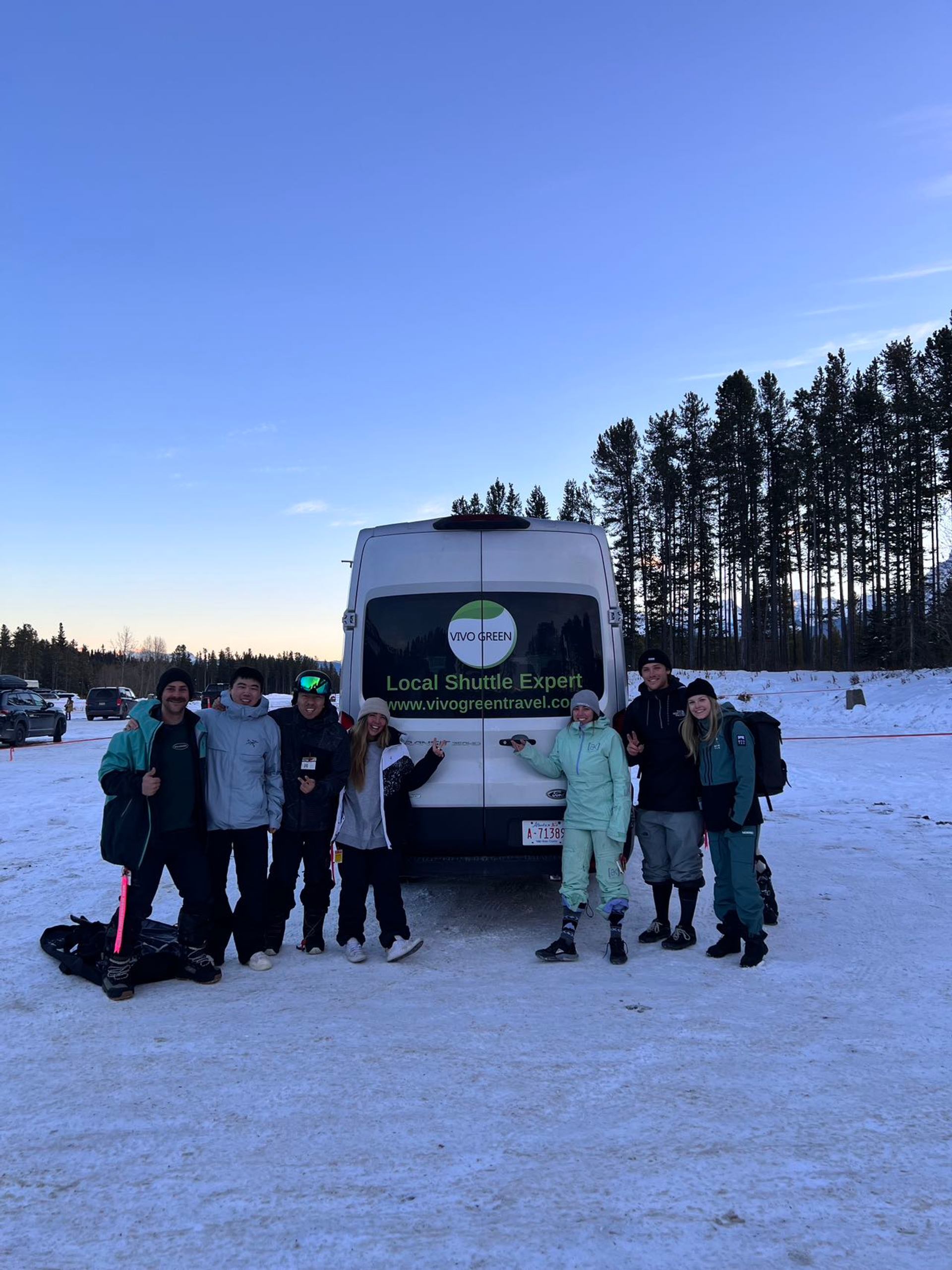 Group in winter clothing stands beside a Vivo Green shuttle van on snow.