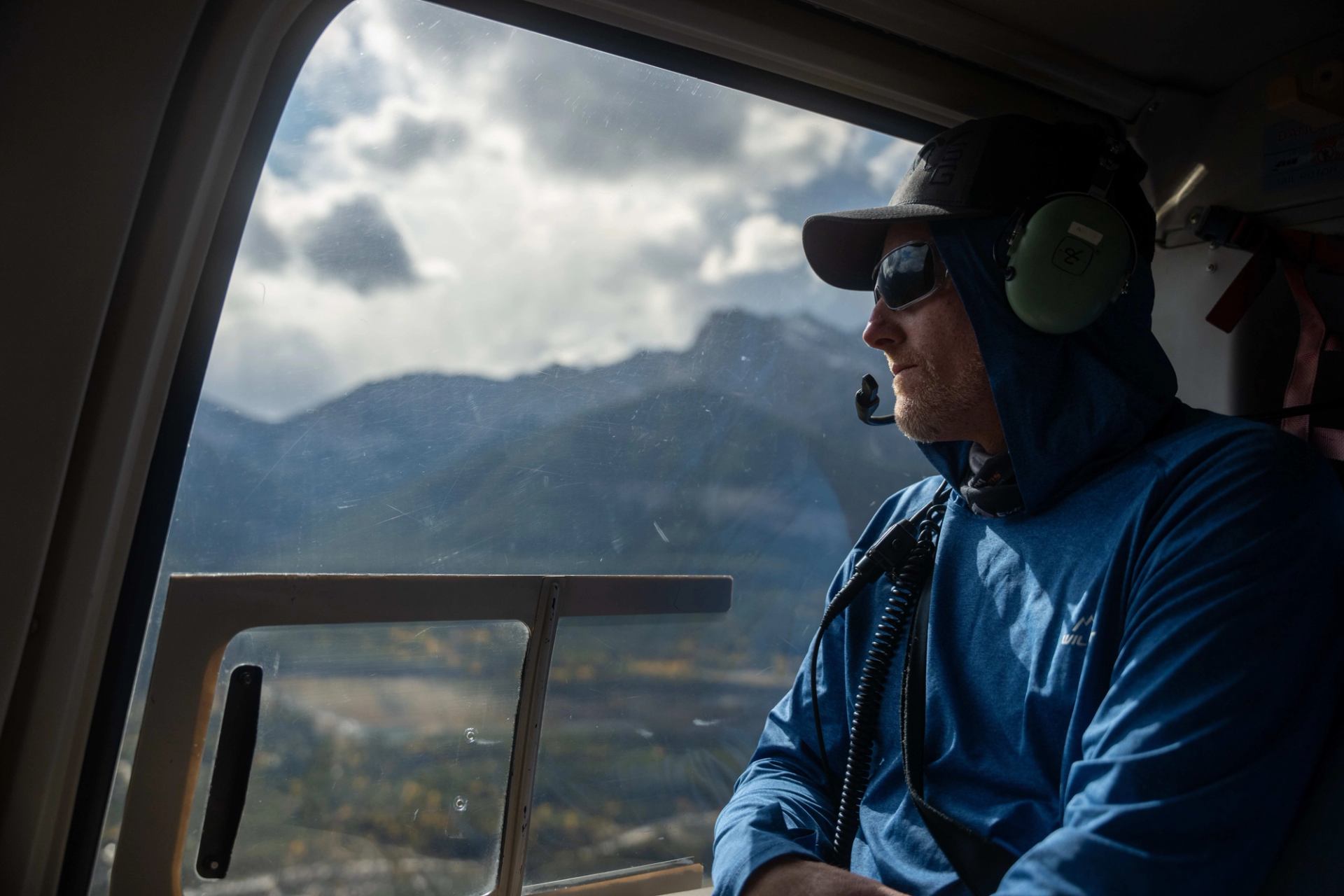 Person in blue jacket gazing at mountain scenery from a helicopter window.