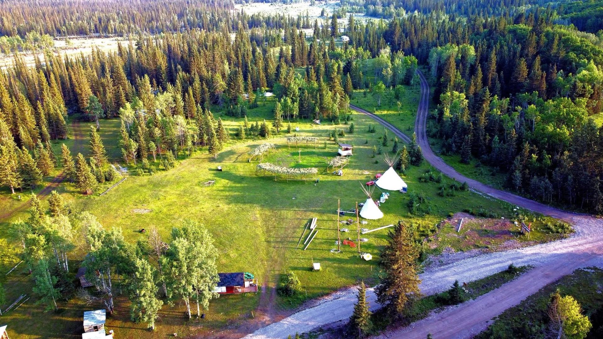 Aerial view of forest clearing with tipis, dirt road and scattered small structures.